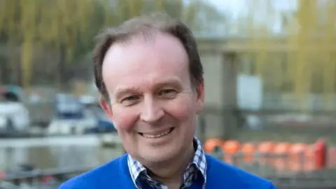 White man in blue T-shirt and check shirt smiles a the camera