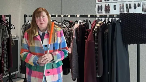 Jane Manley stands in front of Chet Lo's London Fashion Week collection. She wears a colourful checked jacket of greens, creams, blues and pink accompanied by a long orange skirt and a cane with stripes