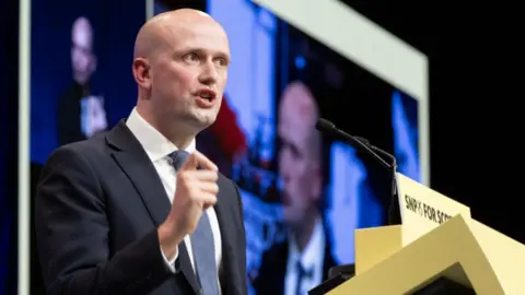 Stephen Flynn, a bald man in a dark blue suit with lighter blue tie, speaks at the podium of the SNP conference 