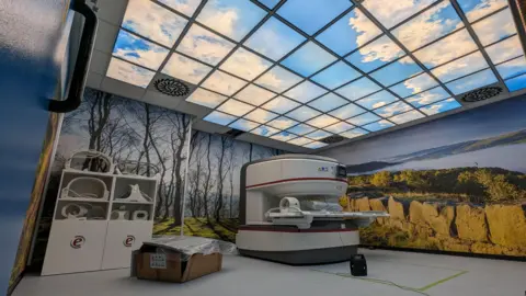 An open MRI scanner in a hospital room with big images of landscapes across two walls and an image of a blue sky and clouds of the ceiling