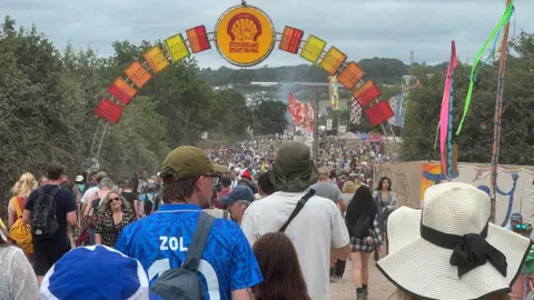 A crowd of people is walking along a dirt road on a festival site. They are wearing hats and T-shirts. There is a gate made of orange and red metal.