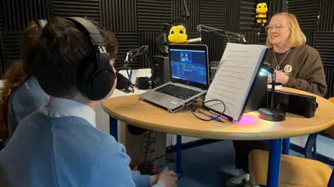 A small group sits around a circular studio table with microphones, headphones, and a laptop running audio or video software. A script stand with printed notes is positioned on the table. The room is lined with acoustic foam panels, and plush bee toys hang on the wall in the background.