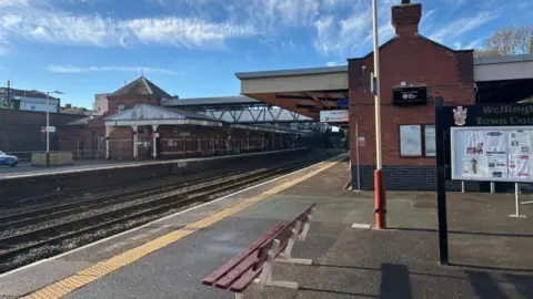 A railway station's platform under a clear blue sky. There is a red and white bench in the foreground with brick railway buildings behind, a thick yellow line on the platform edge and two sets of rails between the platforms.