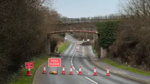 Frank Schuengel A row of red and white cones block off a stretch of road lined with trees, and a road closed sign to the left. Behind is a stone bridge and further roadwork infrastructure behind.