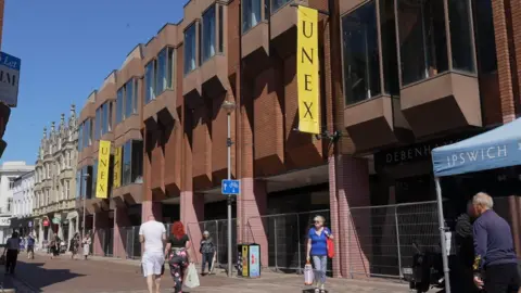 John Fairhall/BBC Westgate Street in Ipswich on a sunny day. The former Debenhams is shown with fencing blocking off its perimeter as shoppers go about their business.