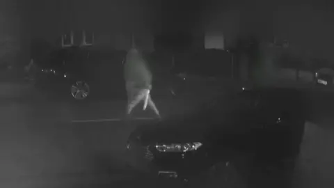 Avon and Somerset Police A black and white screenshot of CCTV footage from a house opposite the street. It shows one of the defendants walking across the road, carrying a large machete which is glinting in the streetlights.