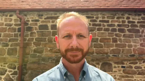 Deputy Tom Coles is wearing a light blue button-down shirt and stands outdoors facing the camera. Behind him is a rustic stone wall with a red-tiled roof and a drainpipe visible on the left side. The image is brightly lit by sunlight, with the background slightly blurred, drawing attention to Tom Coles in the foreground.