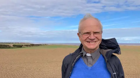 A male priest wearing a black jacket over blue jumper, grey shirt and white dog collar smiles as he stands in front of a brown field that stretches towards the coast, where a strip of grey-blue sea can be seen under a blue sky, with white wind turbines in the distance on the horizon.