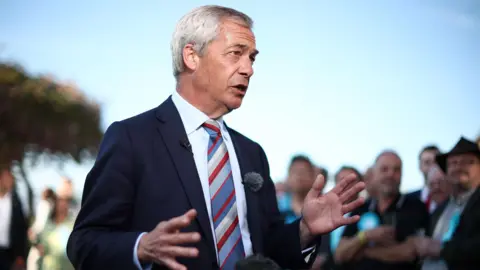 Reform UK leader Nigel Farage attends a post-election event at the Staffordshire County Showground. He is wearing a suit and tie and gesturing with his hands and there are people gathered around him, listening.