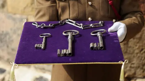 PA Media A purple pillow trimmed with gold, held by a soldier in brown uniform and white gloves, holds three large silver keys and a chain