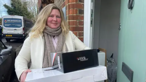 Clare Cowan/BBC Terri Dempsey - a woman with blonde hair, wearing a white coat and grey scarf, holds a white box and a black box