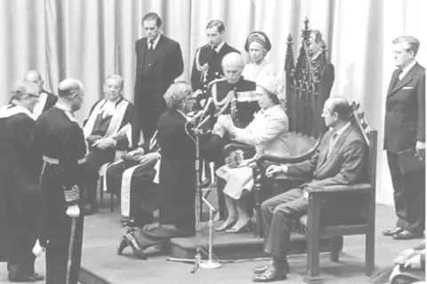 Office of the Lieutenant-Governor A black and white photo of a young Queen Elizabeth II sitting in a ceremonial wooden chair with several people in suits in military uniforms surrounding her