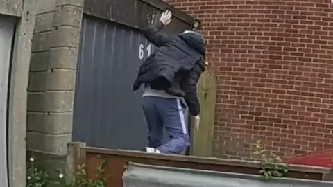 A man in tracksuit bottoms and a padded jacket is seen on the other side of a wooden fence, running away.