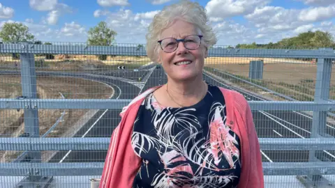 Qays Najm/BBC Carol Charles has white hair and is standing on the new bridge spanning the A47. The road is empty of traffic. Charles wears a black, pink and white frock and a matching pink cardigan. 