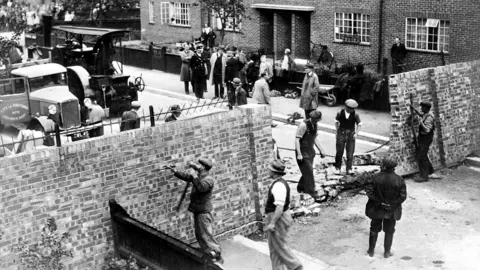 Oxfordshire History Centre Black and white archive image of people knocking down one of the Cutteslowe Walls in 1959. The site is a buzz with workers and vehicles.