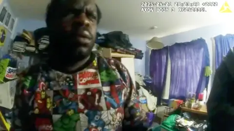 Met Police Police bodycam footage captures Levy being arrested in his bedroom, in April 2025. He is wearing a brightly coloured top. There are purple curtains drawn across the window.