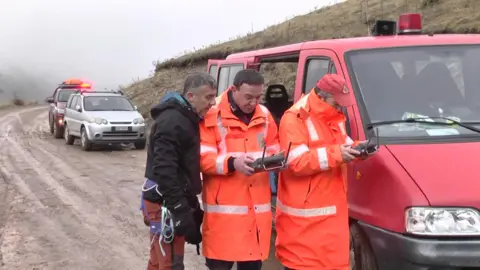 Three men wear orange jackets looking at a handset controlling a drone on a muddy road