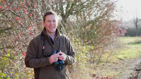 Supplied A man standing in front of bushes with wild red fruit. He has short, brown hair, is wearing a brown fleece jacket, holds black binoculars and he is smiling.