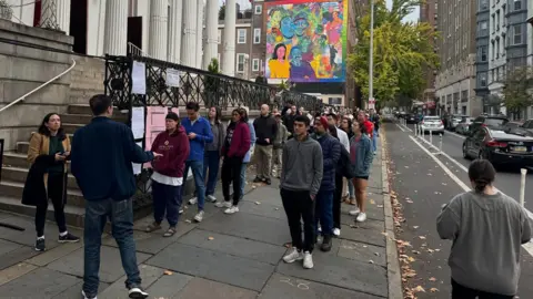 Two rows of people on a sidewalk, most of them wearing dark sweatpants 