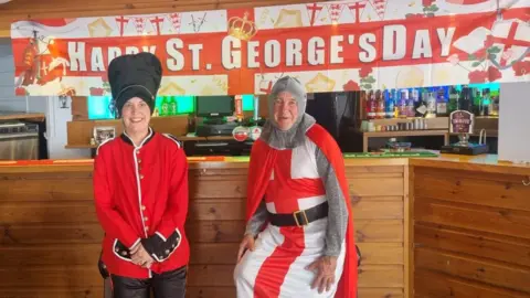 Jerry Kunkler A woman in red Beefeater outfit with her hands clasped and a man in a medieval knights uniform with an English flag and belt. A bar setting and wooden table is behind them both with a flag which says 'Happy St George's Day'
