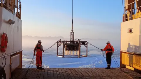 Supplied Two people in orange high-vis jackets and white helmets bringing down a large rectangular contraption using ropes. There are standing on the edge of a platform with the sun and ice sheets in front of them. 