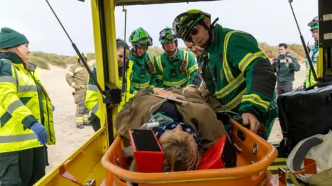 Getty Images A person is lying down in a vehicle while being attended to by workers in hi-vis clothing. In the background there is sand