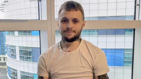 A man with short brown hair, a beard and wearing a white T-shirt sits in front of a window and smiles.
