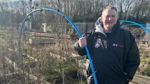 Mark Eyres is standing in front of the overgrown flower beds at the allotment in a black Leicester Tigers overcoat. Behind mark are dark brown garden sheds.