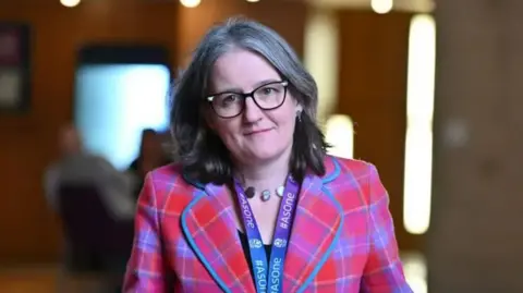 Getty Images Maree Todd looking right at the camera. She has dark hair. She is wearing dark-rimmed glasses and a blue and purple lanyard around her neck. She is wearing a pink tartan jacket with blue and yellow lines.