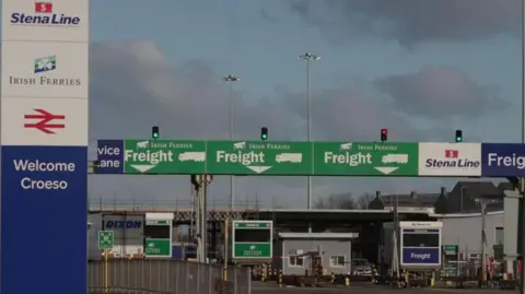 BBC Three green signs above different lanes directing freight traffic on to Irish Ferries service to Dublin are seen at the Port of Holyhead in Holyhead. To the left of these is a large blue and white 'Welcome' sign with the various transport companies listed. There are no vehicles driving towards the barriers.