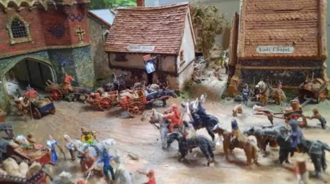 Phil Roberts A model showing the medieval shrine dedicated to Mary in Ipswich. It shows a roadway leading to a gate on the far left, with a two storey tavern building and Lady Chapel on the far side of the road. Models showing people on horseback, pushing carts, on foot and driving carts are moving along the street.