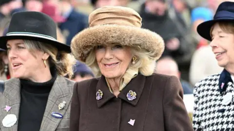 Reuters Queen Camilla, wearing a brown coat and brown hat, smiling towards the camera. She is in between two other women who are wearing hats and smiling. 