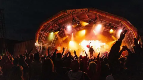 Wychwood Festival A man stands on a stage and lifts up his guitar as he basks in orange light and fans watching on throw their arms up in the air. It is dark. There is a sign that reads "Wychwood" at the top of the stage.