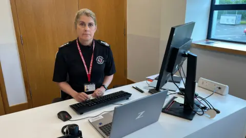 Steve Hubbard/BBC Northamptonshire Fire and Rescue Service chief fire officer Nikki Watson sitting behind a desk. She is wearing a black blouse and has an identity card on a red lanyard around her neck. There is a laptop, monitor and keyboard on the desk.