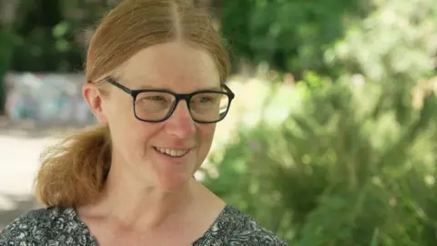 Dr de St Croix is seated on a bench in a garden on a sunny day. She is smiling and has shoulder length hair, which is tied back, and wears glasses. 