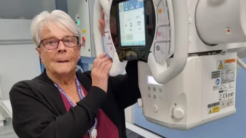 Jan Cottingham looks at the camera and has her hands on a piece of radiography equipment. She has short pale grey hair and is wearing glasses.