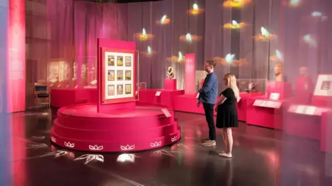 The Trustees of the British Museum The image shows one of the sections of the exhibition, lit in vibrant pink, with visitors seeing the displays.