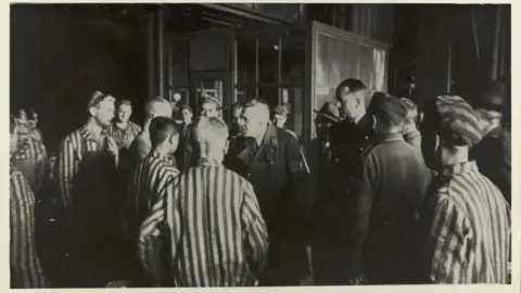 The Wiener Holocaust Library A historical black and white photograph depicting a group of people in striped concentration camp uniforms standing indoors, with a senior Nazi official in a dark uniform speaking to them.