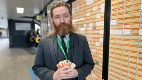 Kate Bradbrook/BBC Elliott Badger with brown hair and a beard wearing a grey suit and a green lanyard holding a stack of orange rail tickets at Northampton railway station in front covered in orange train tickets