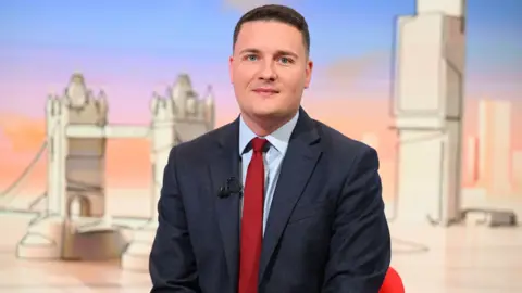 BBC Mid shot of Health Secretary Wes Streeting sitting down in a red chair in some kind of studio with an artistic line drawing representation of Tower Bridge and some other buildings in the background. Mr Streeting is wearing a navy suit with a red tie and light blue shirt.
