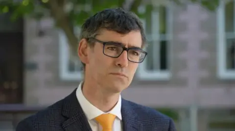 Deputy Montford Tadier is looking serious as he looks at the camera. He is standing outside a pink brick building and is wearing a blue-checked jacket, a white shirt and a yellow tie. He has dark hair and is wearing glasses.