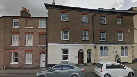 A Google Maps picture of the town council's headquarters, a 19th Century, three-storey, mid-terrace building with two cars parked on the road outside. 