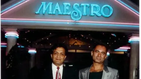 Moss Sheikh/Mick Chandsoor/Legacy Bhangra Daytimers Two South Asian men wearing smart suits stand in front of a building with a blue neon lit sign saying The Maestro.