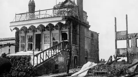 Getty Images The remains of the Royal Box at Hurst Park grandstand in Molesey after it was burnt down in a protest by suffragettes. A man in a suit and a straw boater stands in front of the badly damaged building.