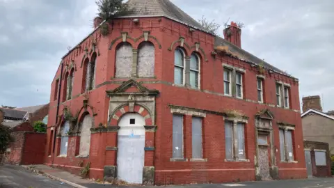 The former Queens Arms. It is a two-storey red-brick property on a street corner. Its doorways and windows are boarded up. Weeds are growing in several spots including through the roof.