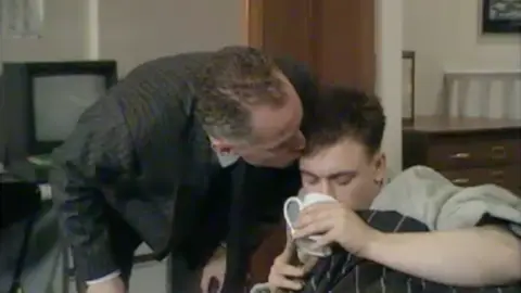 Lord Michael Cashman (left) kissed Gary Hailes (right) on the side of the head. A man wearing a dark suit kisses a man in a grey jumper holding a cup.