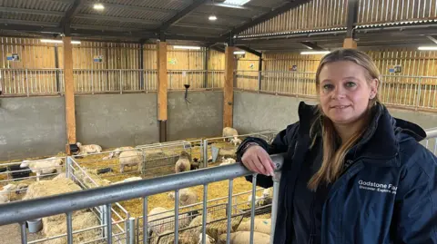 BBC / Adrian Harms A woman with light brown hair leans on a fence on a mezzanine. Below her are sheep on the ground floor.