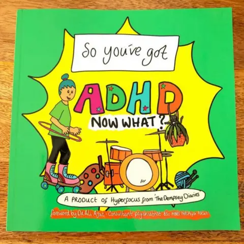 Bethany Dempsey A picture of a green book on a wooden table. The title reads "So You've Got ADHD, Now What?" The text underneath reads: "A product of hyperfocus from The Dempsey Diaries. The illustration on the front is of a woman in a green top with a hula hoop around her waist and wearing roller skates standing next to a drum kit, with knitting on the floor. The illustration is in bright primary colours."