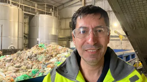 Brazilian man wearing high viz and protective glasses smiles at camera, behind him is a skip full of food waste bags piled up and some large silver tanks.