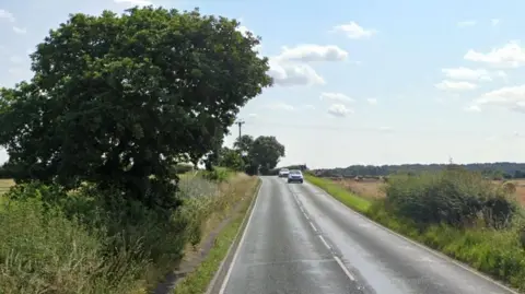 A single carriageway road with two cars in the distance. There is grass and a tree on the left and grass and fields on the right.